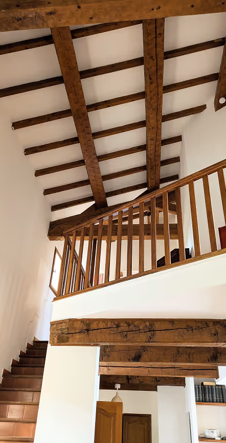 Intérieur d'une pièce avec poutres en bois au plafond, escalier en bois et rambarde en bois surplombant un étage supérieur.
