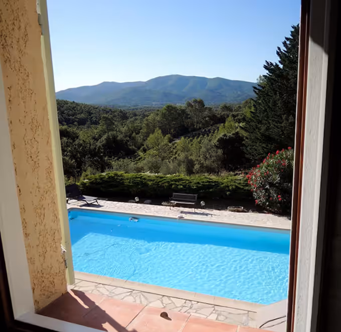 Vue depuis une fenêtre sur une piscine bleu vif et sa terrasse, avec arbres verts et montagnes en arrière-plan sous un ciel dégagé.