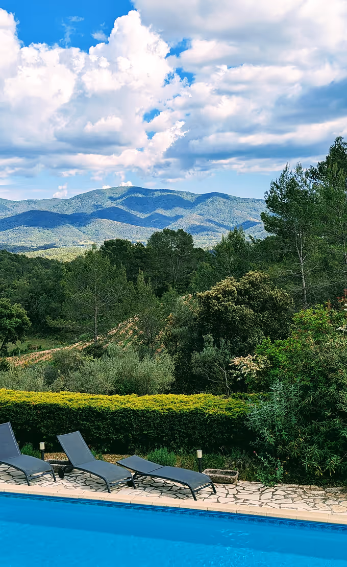 Piscine bleue avec trois chaises longues noires sur une terrasse en pierre, haies, arbres verts denses et montagnes bleues au loin sous un ciel partiellement nuageux.