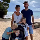 Famille souriante de cinq personnes posant sur une plage, avec des arbres et un ciel nuageux en arrière-plan.