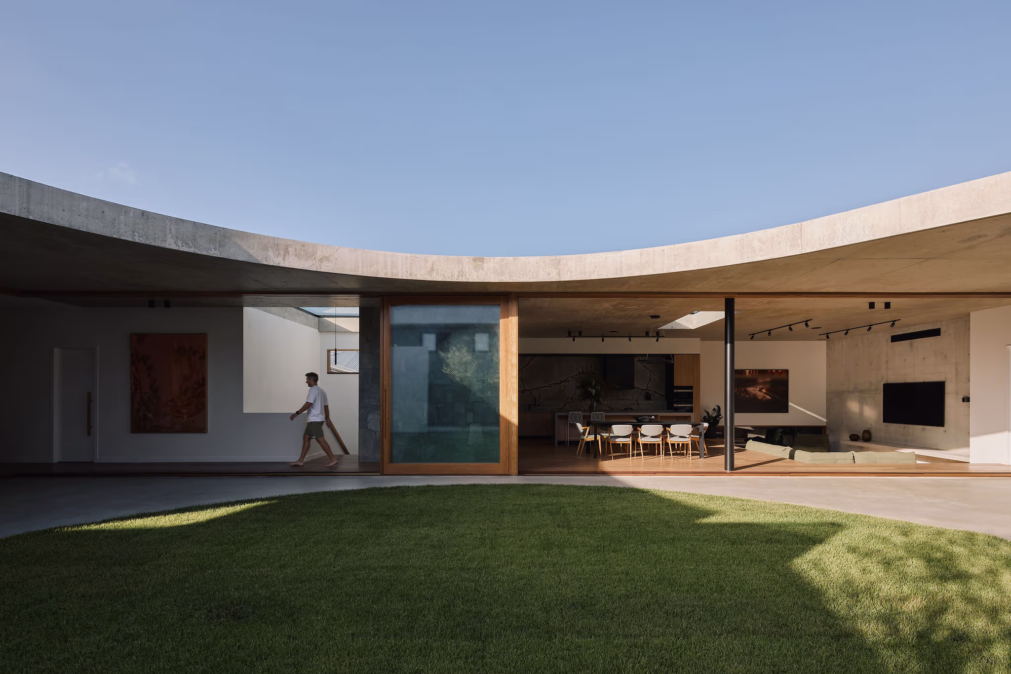 Modern courtyard house with open living and dining area, featuring a person walking inside under a curved concrete roof.