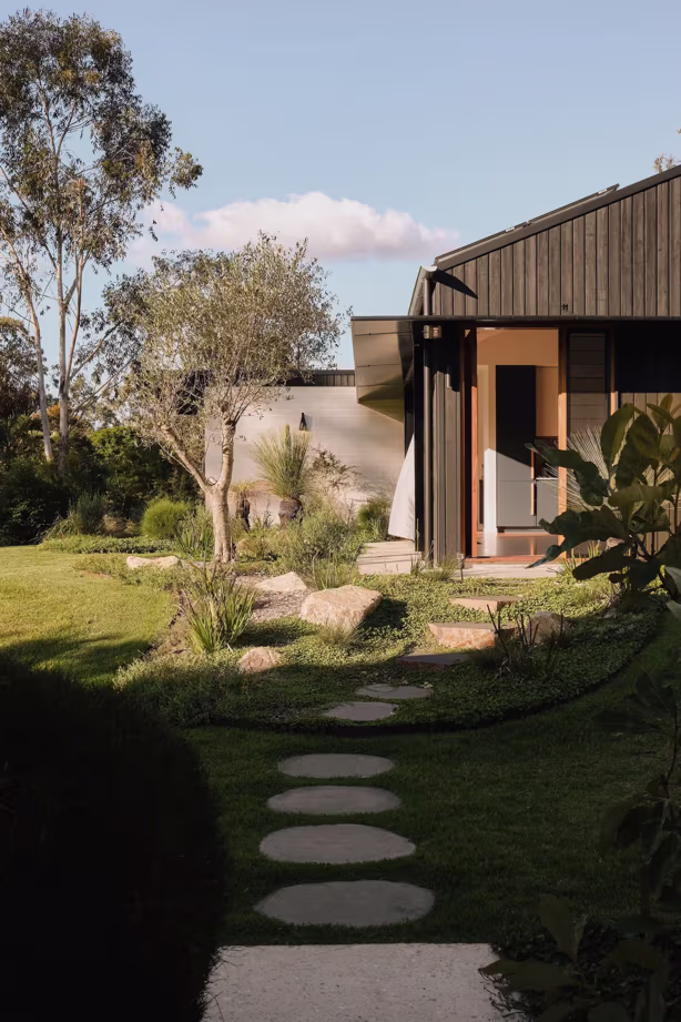 Modern house entrance with a stone pathway leading through a green garden and trees under a blue sky.