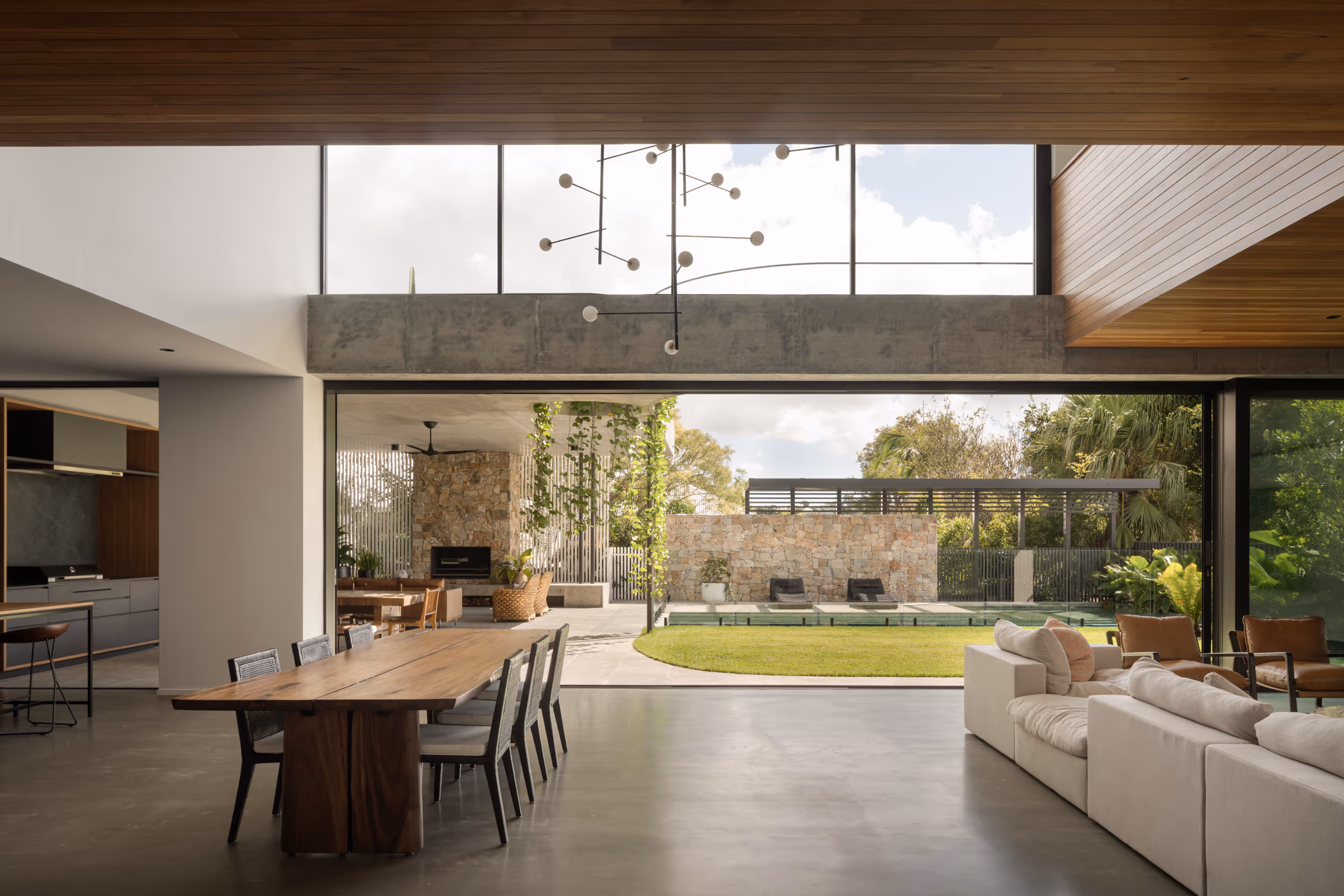 Modern open-plan living room and dining area with large wooden table, beige sofa, and view of a garden and pool through large glass doors.