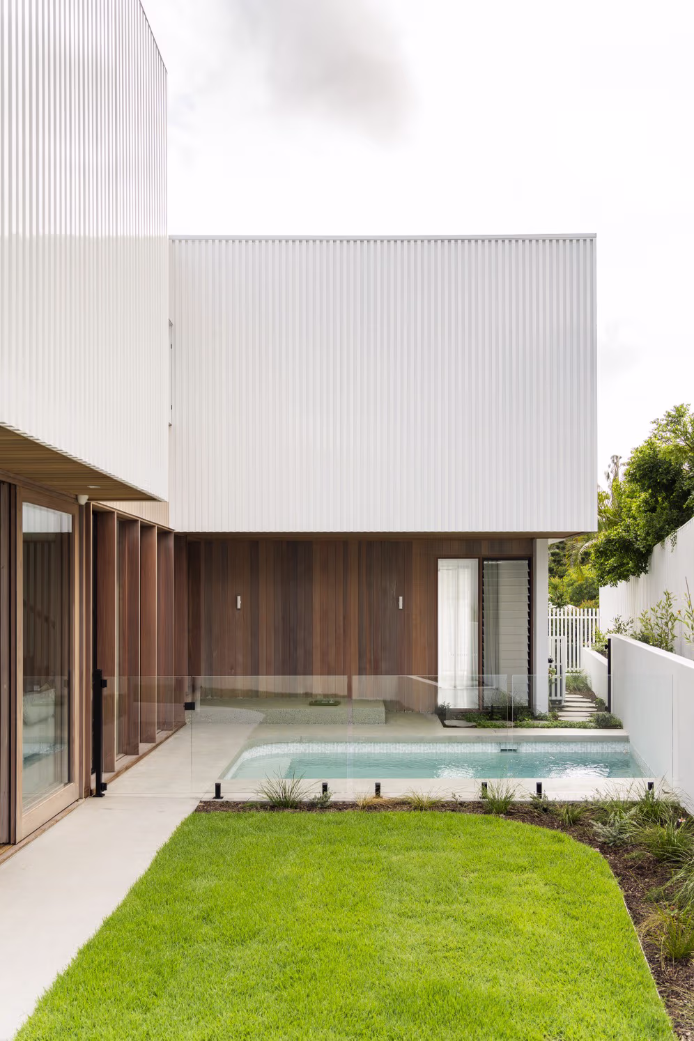 Modern house backyard with a small rectangular pool, glass fence, wooden paneling, and bright green lawn.