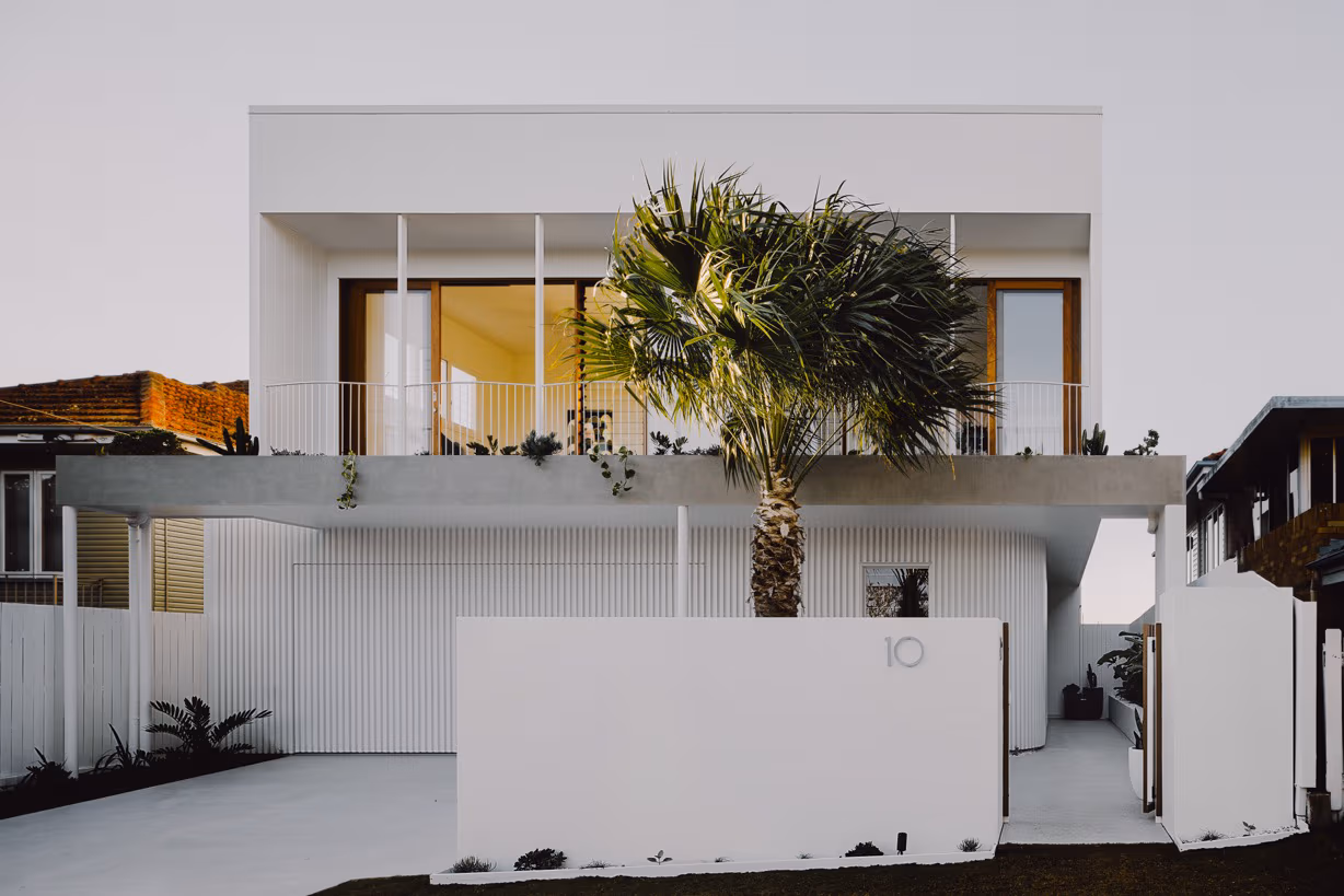 Modern two-story white house with large balcony, central palm tree, and fenced front yard.