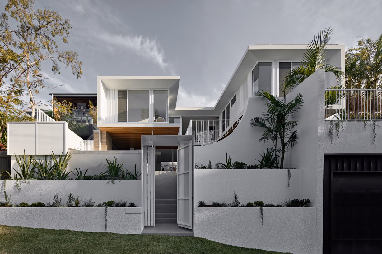Modern white house with large windows, curved walls, and tropical plants in front under a cloudy sky.
