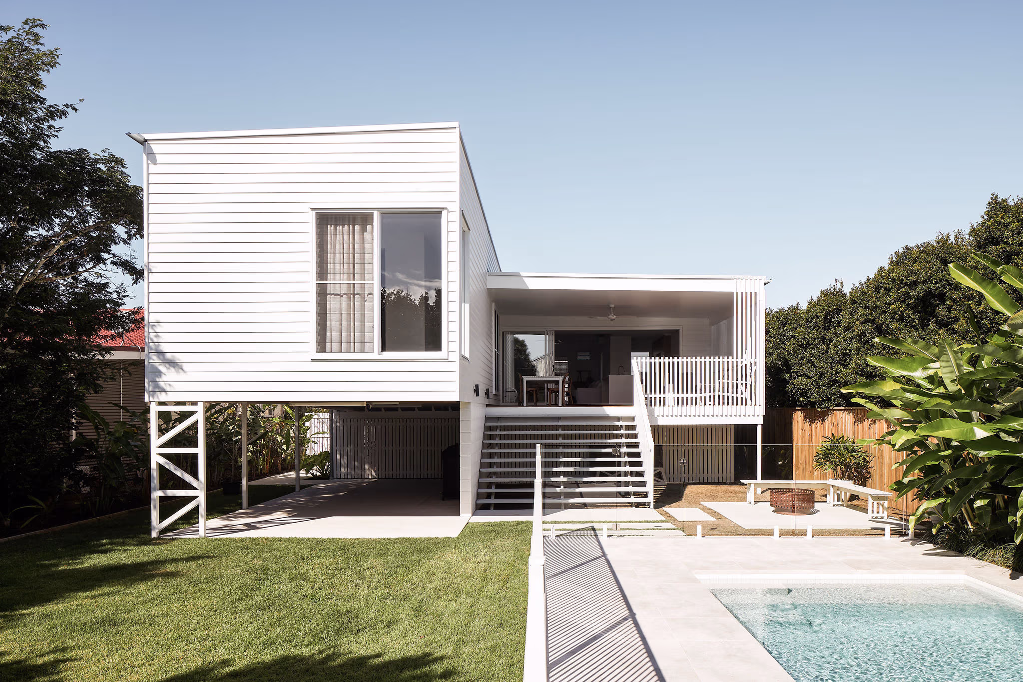 Modern white elevated house with outdoor stairs, patio furniture, and a pool in the foreground surrounded by green lawn and plants.