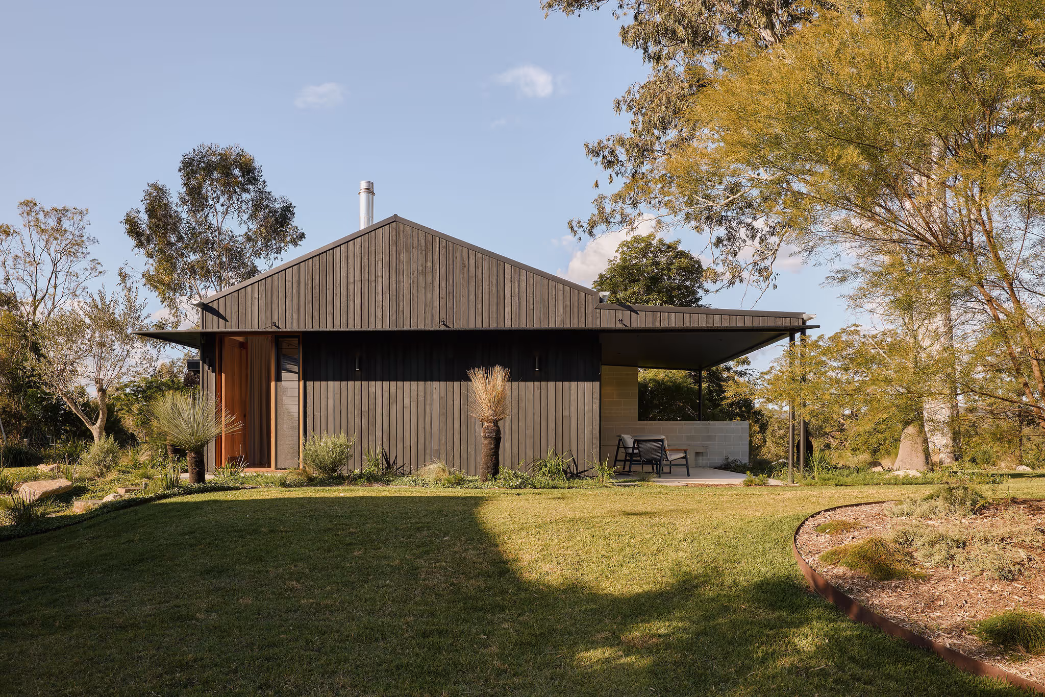 Modern single-story house with vertical dark wood siding, a small front porch with chairs, surrounded by grassy lawn and trees.