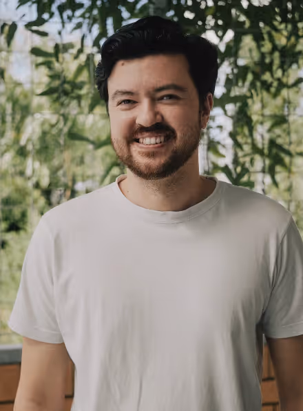 Smiling man with dark hair and beard wearing a plain white t-shirt standing in front of green leafy plants.