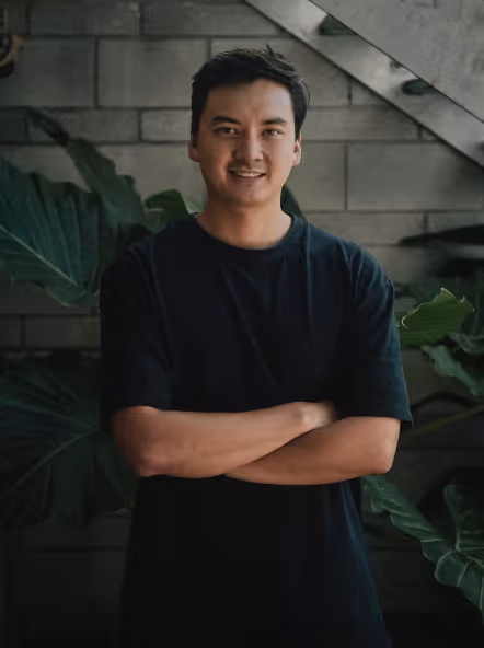 Smiling man in a black t-shirt standing with arms crossed in front of large green leaves and a stone wall.