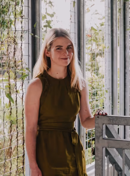 Woman with long blonde hair wearing an olive green sleeveless dress, standing by a metal railing in a plant-filled greenhouse.