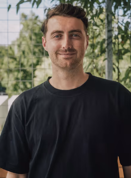 Smiling man with short brown hair wearing a black t-shirt standing outdoors with greenery in the background.