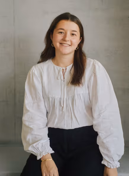 Smiling woman with long dark hair wearing a white blouse and black pants sitting against a plain gray background.