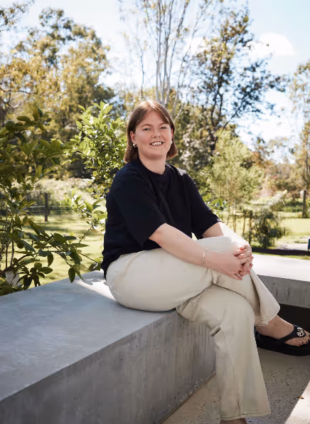 Woman with short brown hair wearing a black shirt and beige pants sitting on a concrete bench outdoors with greenery in the background.
