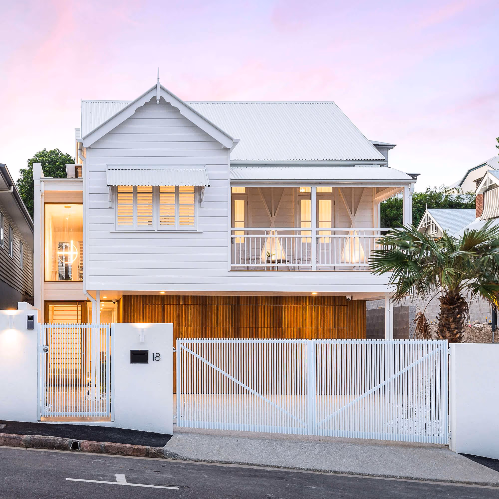 Modern two-story white house with a balcony, wooden garage door, and white fence with gate at dusk.