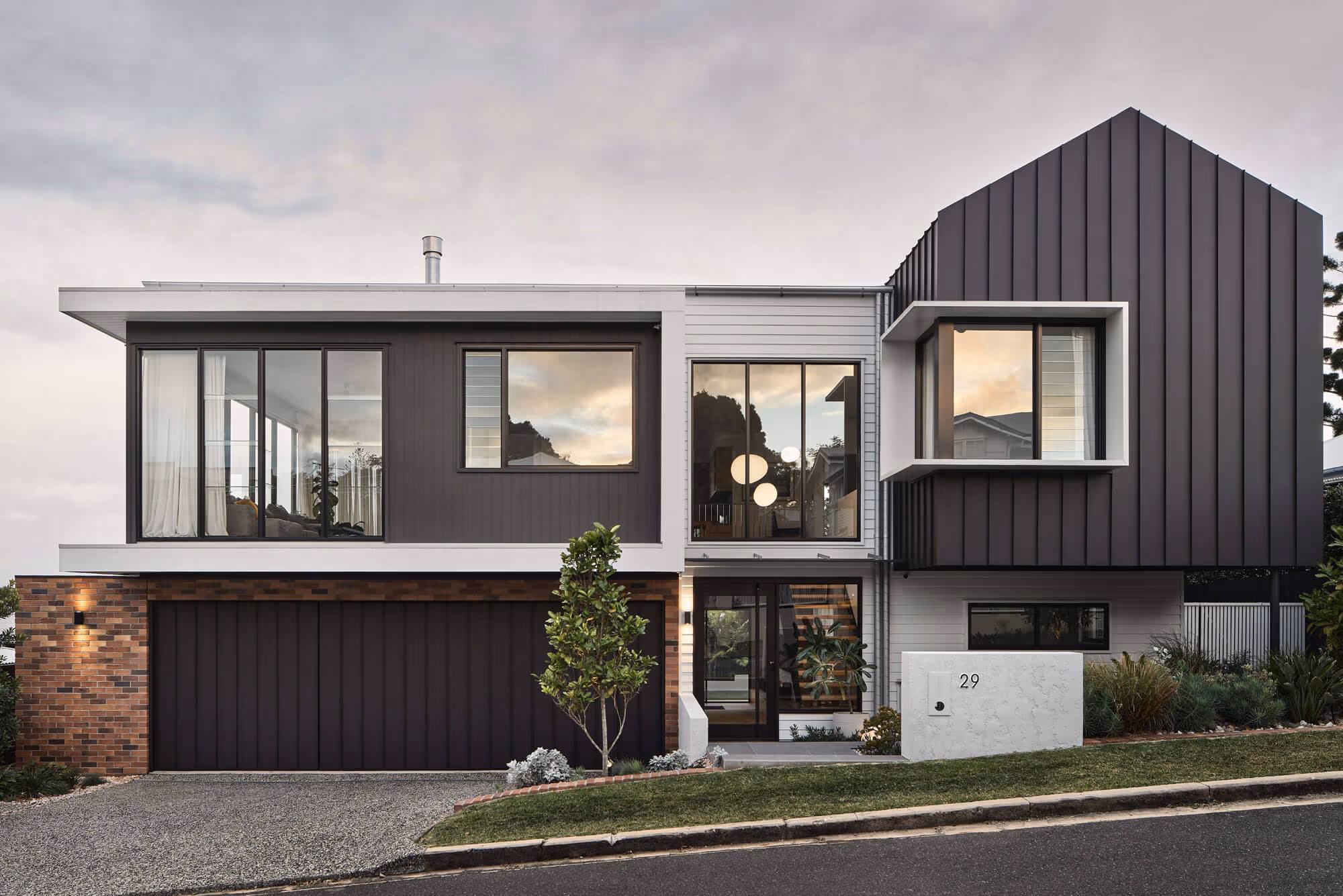 Modern two-story house with black vertical siding, large windows, brick garage, and landscaped front yard at dusk.