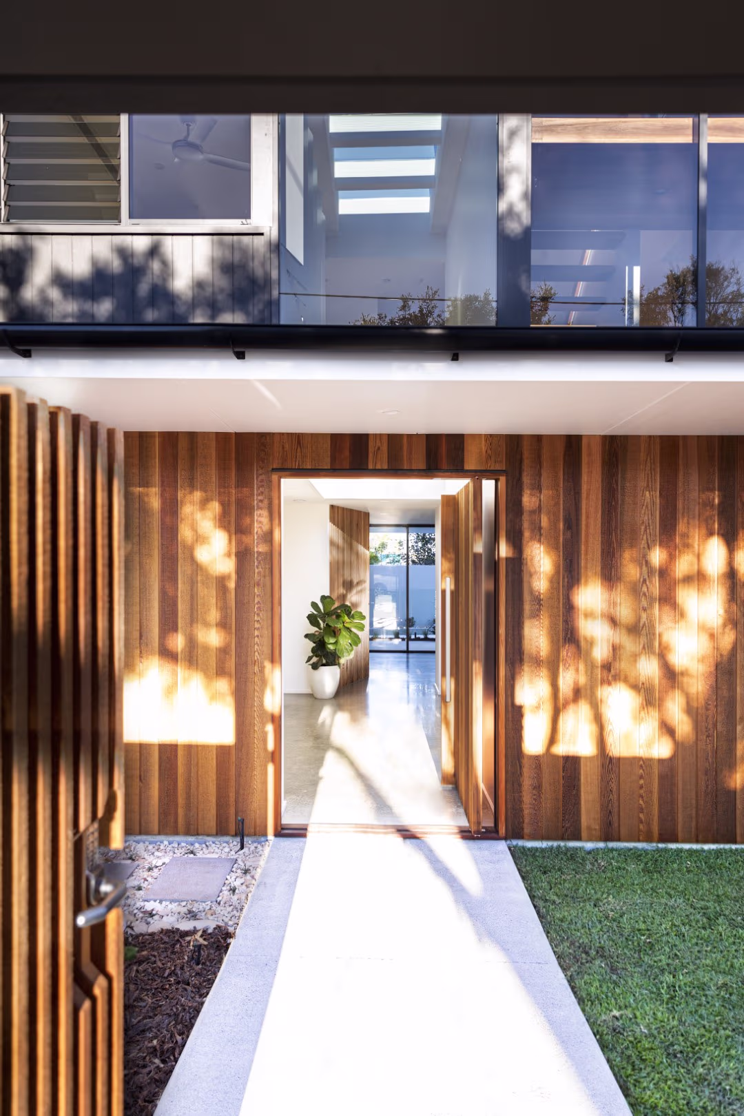 Modern house entrance with wooden paneled walls, a large open wooden door, a concrete pathway, and a potted plant inside.