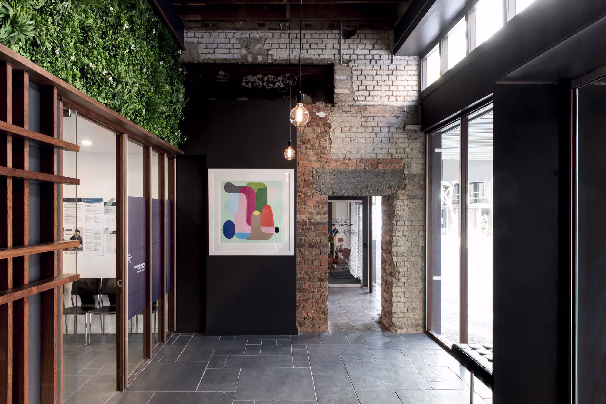 Modern interior hallway with exposed brick wall, black walls, gray tiled floor, a colorful abstract painting, and hanging light bulbs.