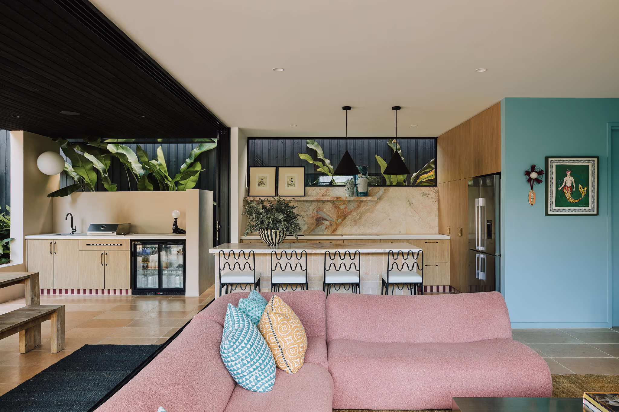 Modern open living room with a pink sectional sofa, colorful patterned pillows, and a kitchen area with bar stools, plants, and artwork.