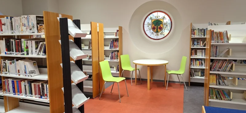 Coin de la bibliothèque avec des étagères en bois remplies de livres, trois chaises vertes autour d'une table ronde blanche sous une fenêtre circulaire avec un vitrail décoratif.