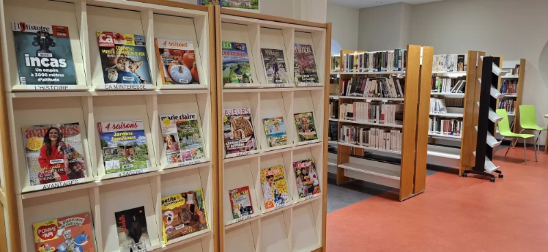 Rayonnage en bois avec des magazines d'actualité et de loisirs exposés dans la bibliothèque, avec d'autres étagères contenant des livres en arrière-plan et quelques chaises vertes près du mur.
