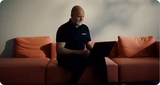 En mann med skjegg sitter på en oransje sofa og arbeider på en bærbar datamaskin.