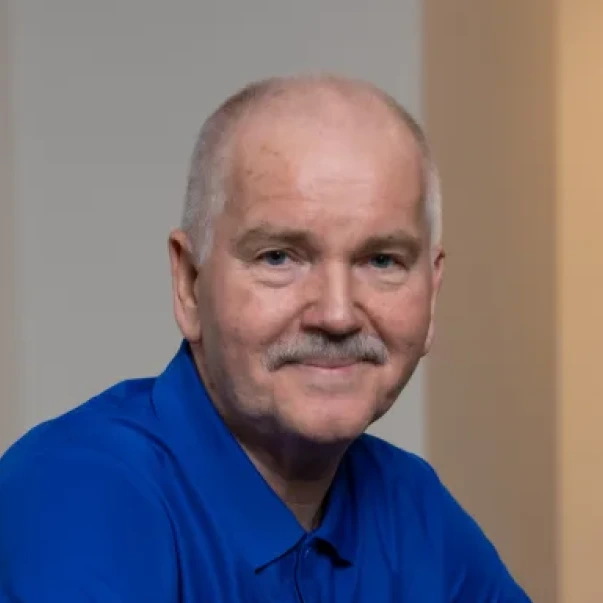 Portrett av en eldre mann med blå skjorte som smiler svakt i et innendørs miljø.