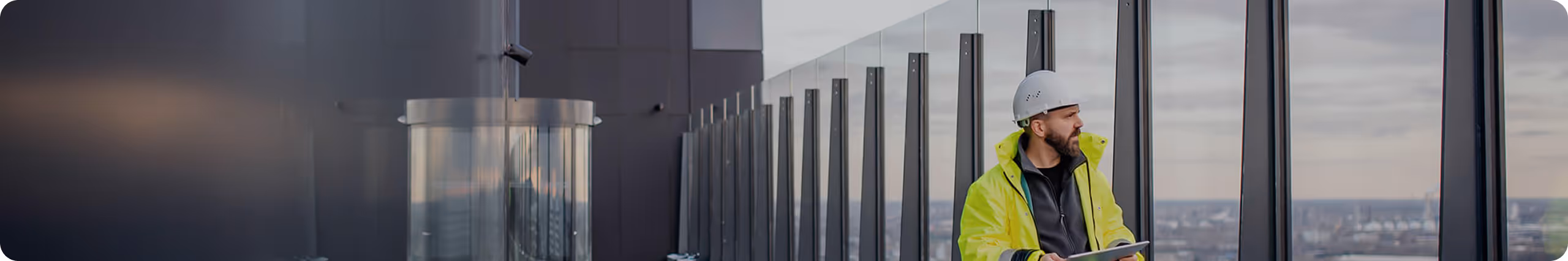 Bygningsarbeider i gul sikkerhetsjakke og hjelm som ser ut over bylandskap og holder et nettbrett ved en glassrekkverk på et høyt bygg.