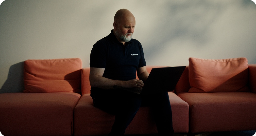 Mann med skjegg som sitter på en oransje sofa i dempet lys og bruker en bærbar PC.