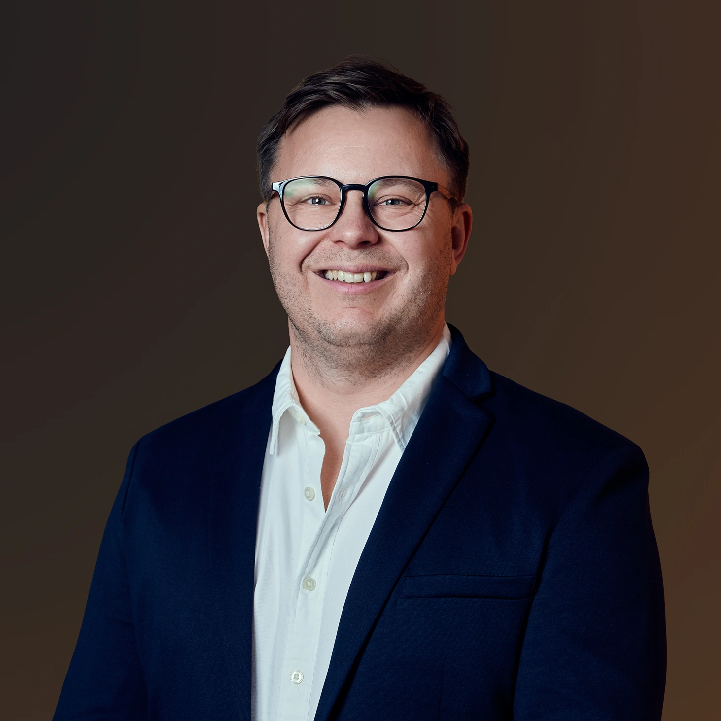 Mann med briller smiler, iført hvit skjorte og mørk blazer foran en ensfarget bakgrunn.