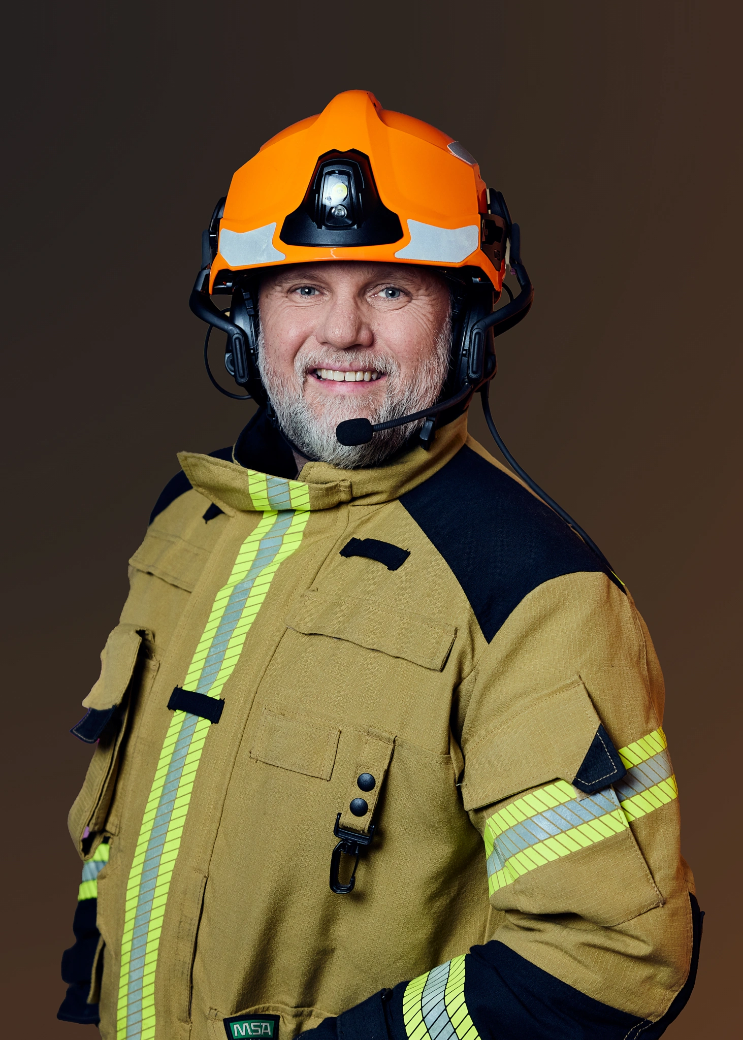 Portrett av en smilende brannmann med grått skjegg iført oransje hjelm og gul refleksjakke.