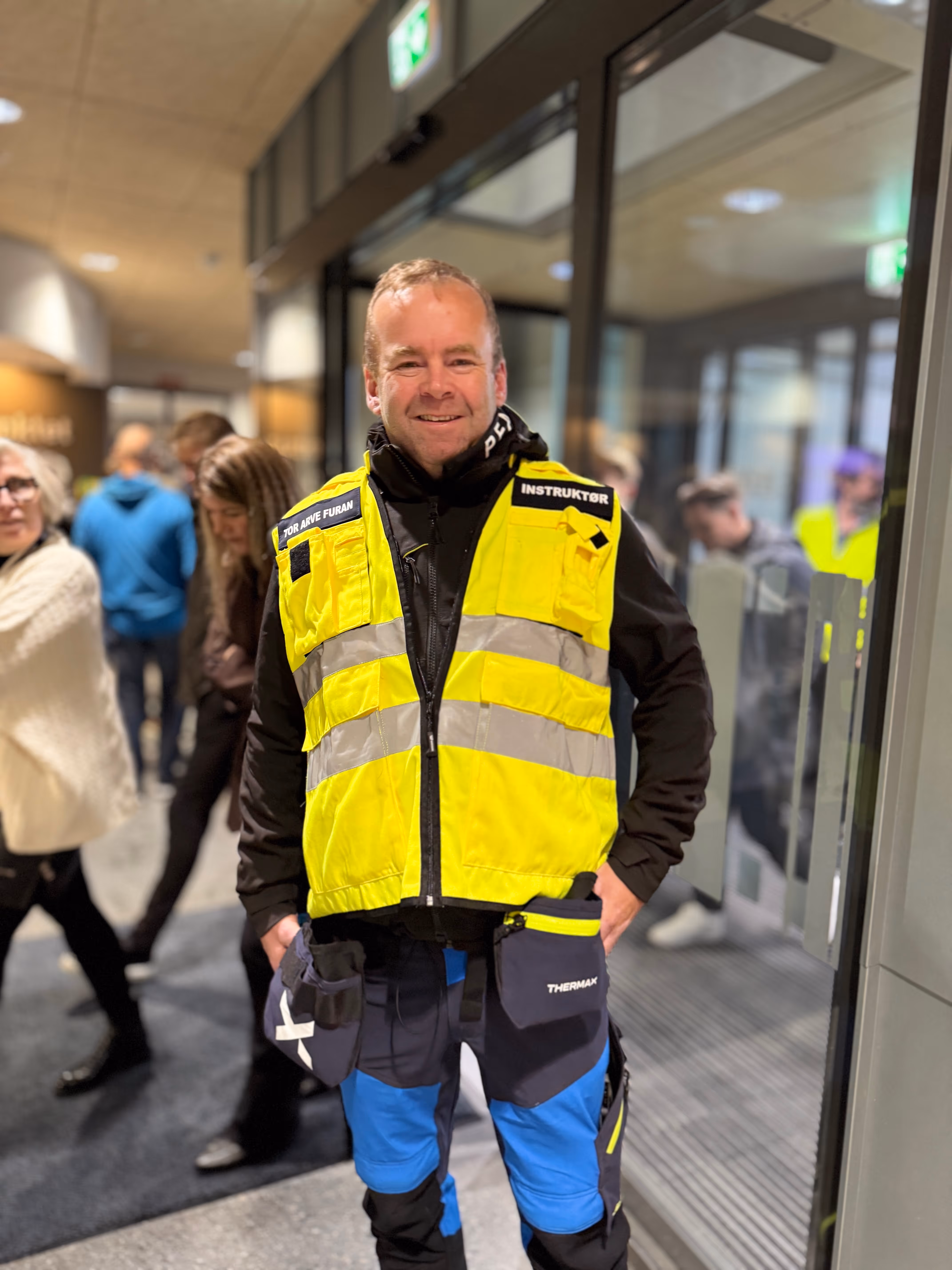 Mann med gul refleksvest merket 'INSTRUKTØR' og arbeidsbukser, står innendørs foran en glassdør med folk i bakgrunnen.