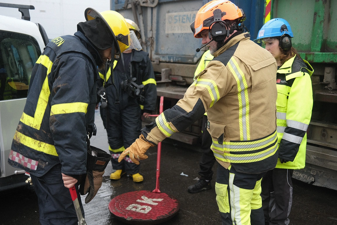 Brannmenn og arbeidere i verneutstyr og hjelmer samles rundt en rød kumlokk merket BK på en våt gate.