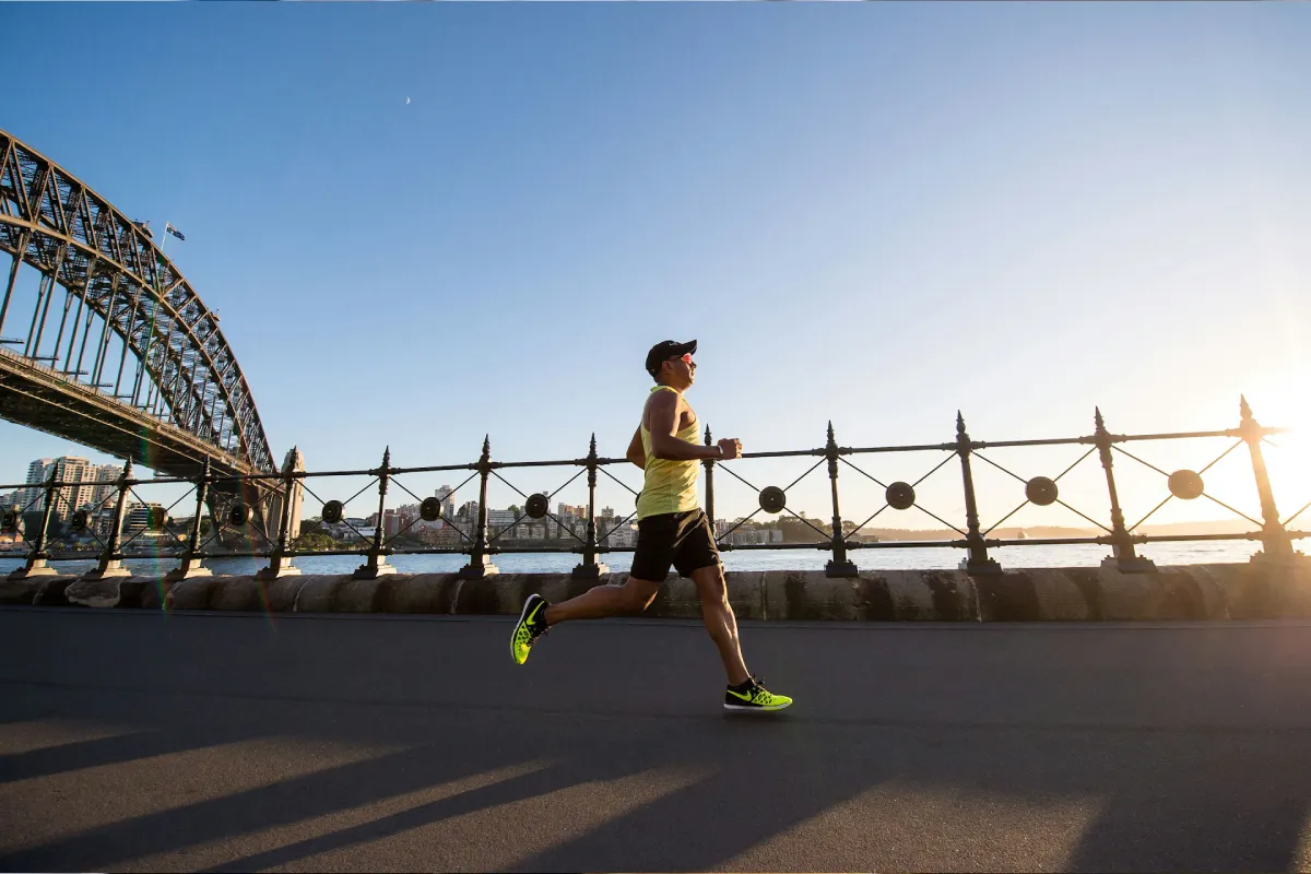 Man jogging along waterfront near a metal fence with a large arched bridge and city skyline in the background at sunset.