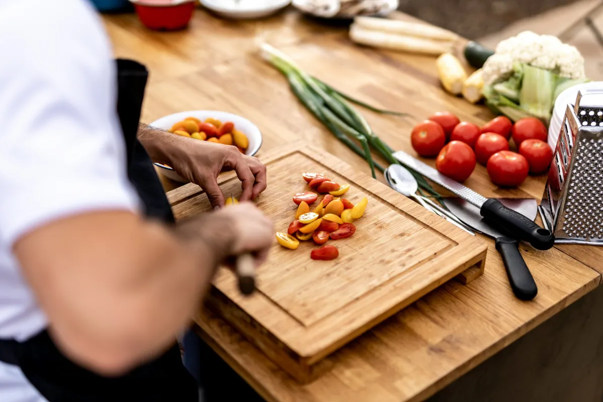 Person slicing red and yellow cherry tomatoes on a wooden cutting board in a kitchen with fresh vegetables nearby.