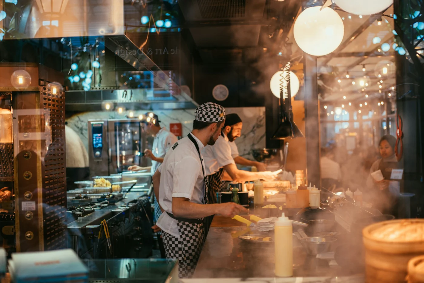 Chefs cooking and preparing food in a busy restaurant kitchen filled with steam and warm lighting.