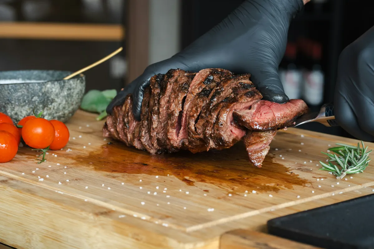 Chef wearing black gloves slicing grilled steak on a wooden cutting board with cherry tomatoes and rosemary nearby.