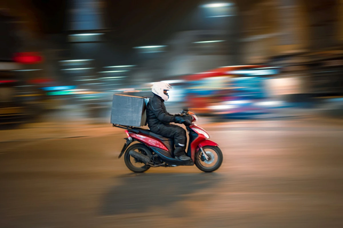 Delivery person wearing a white helmet riding a red scooter at night with a large box on the back.