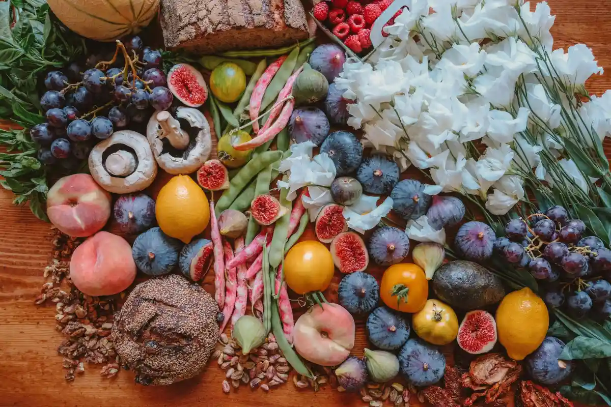 Assortment of fresh fruits, vegetables, nuts, mushrooms, bread, and white flowers arranged on a wooden surface.