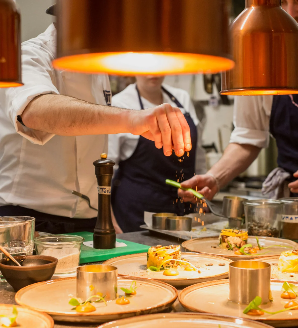 Chef sprinkling seasoning over plated gourmet dishes in a busy kitchen.