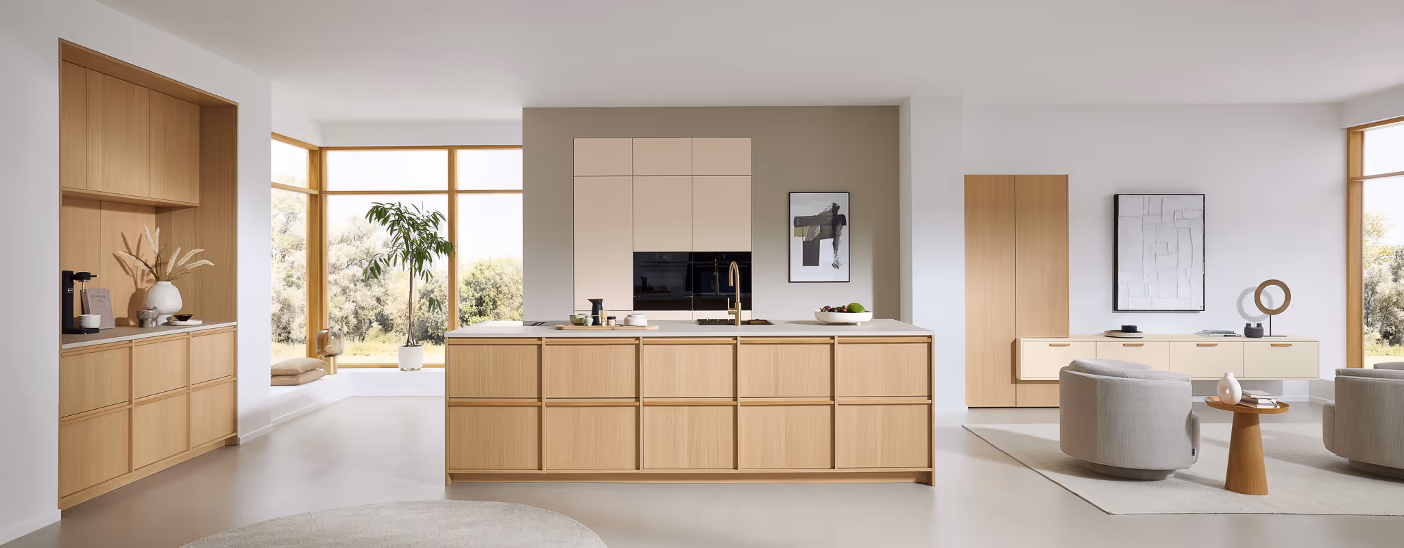 Moderne Küche mit hellen Holzmöbeln, Kücheninsel, großen Fenstern und angrenzendem Sitzbereich mit zwei grauen Sesseln.