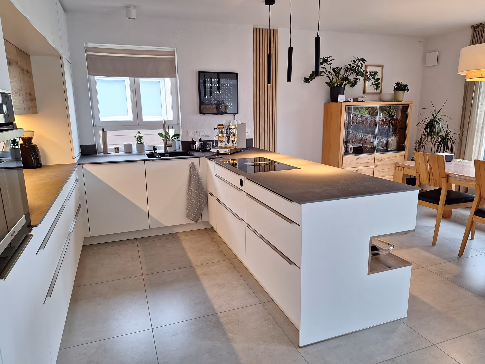 Moderne weiße Küche mit Kücheninsel, Induktionskochfeld, Kaffeemaschine und Holzschrank neben Essbereich.