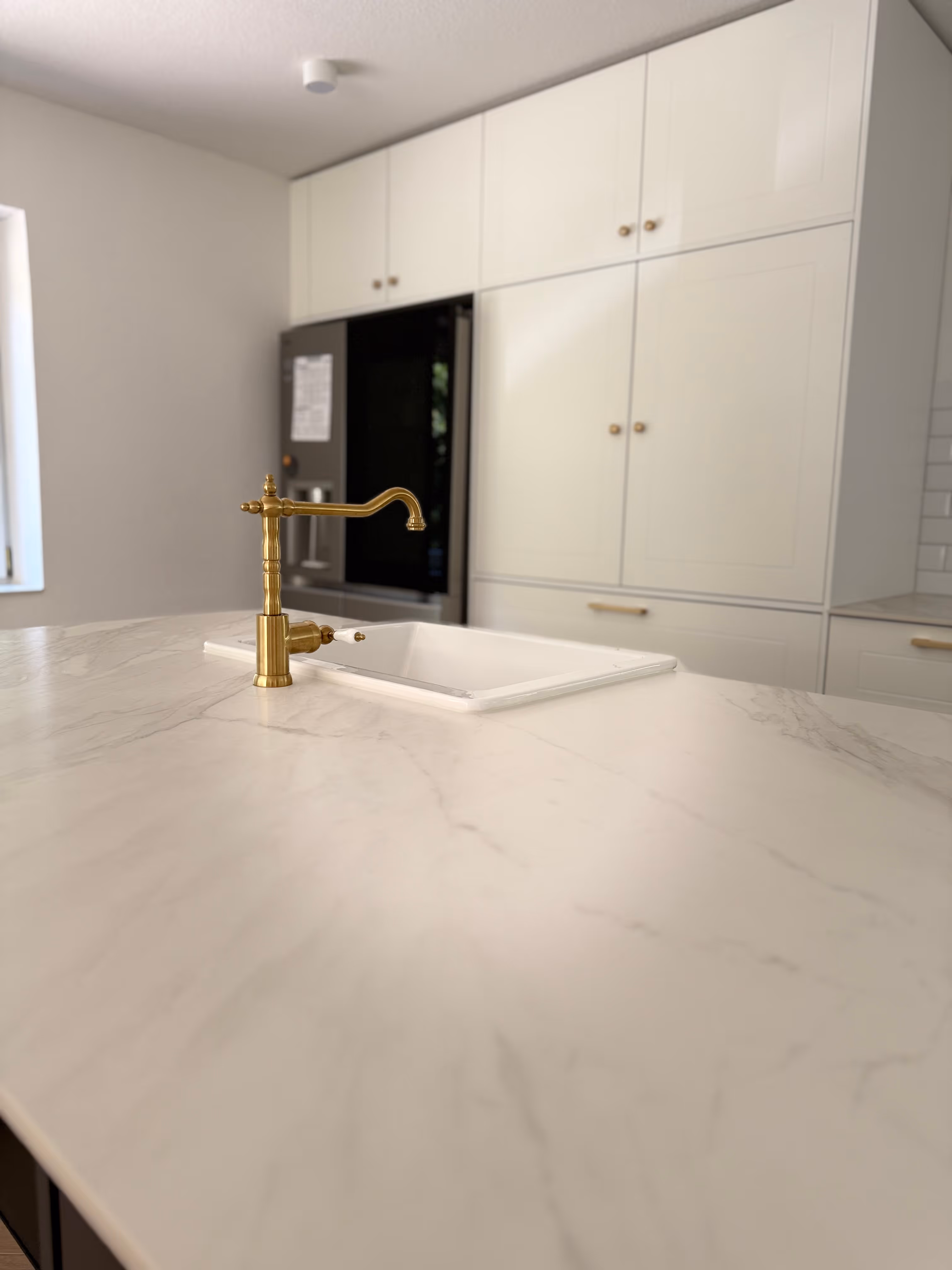 Moderne Küche mit weißer Marmor-Arbeitsplatte, goldfarbenem Wasserhahn und eingebautem Spülbecken vor weißen Schränken.