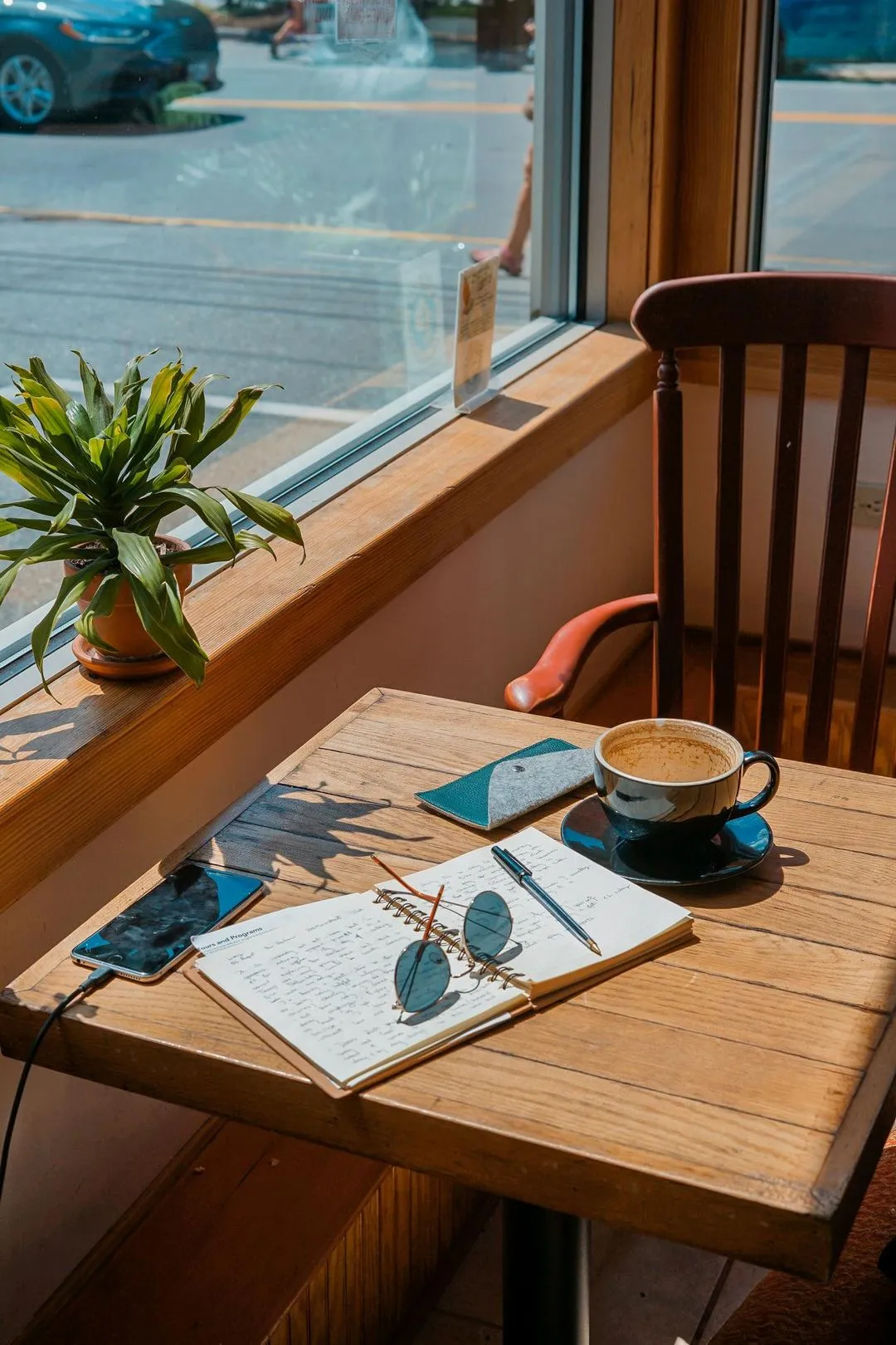 Cozy cafe scene with a wooden table by a sunlit window. On the table are a notebook, sunglasses, a pen, a smartphone, and a cup of coffee. A potted plant adds a fresh touch.
