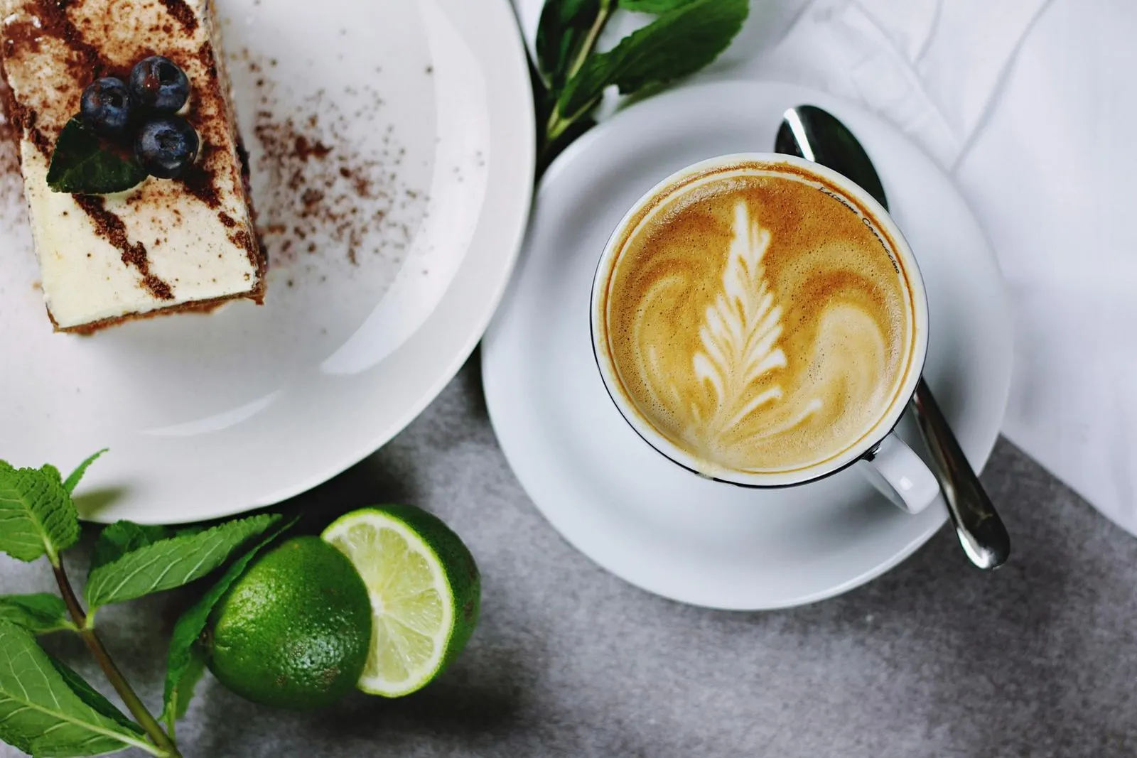 A latte art coffee cup beside a slice of cake topped with blueberries, garnished with mint and limes on a gray table.