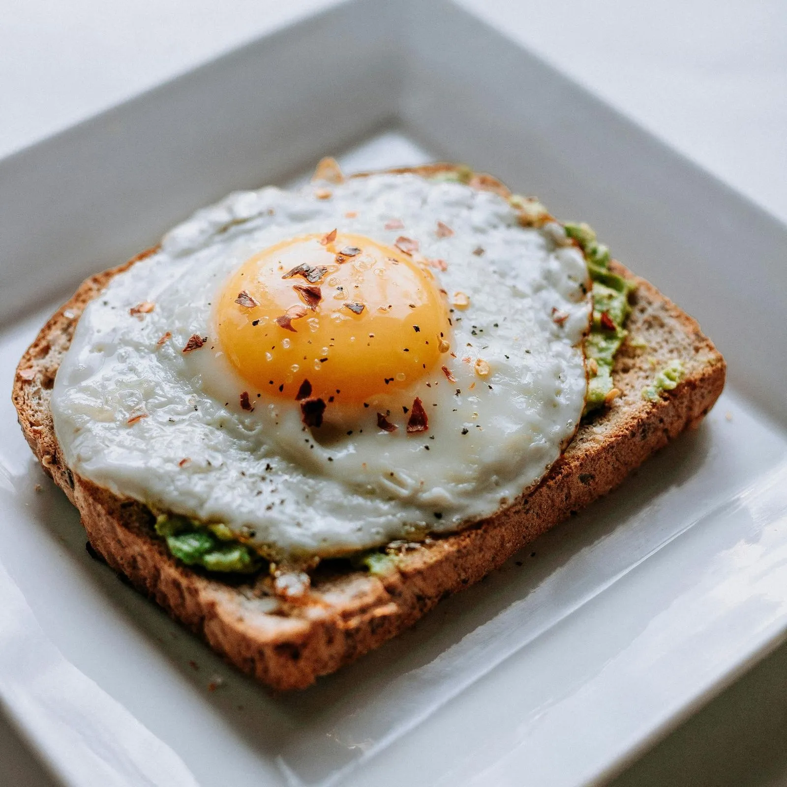 A slice of whole grain toast topped with a perfectly fried egg, featuring a bright yolk and sprinkled with chili flakes.