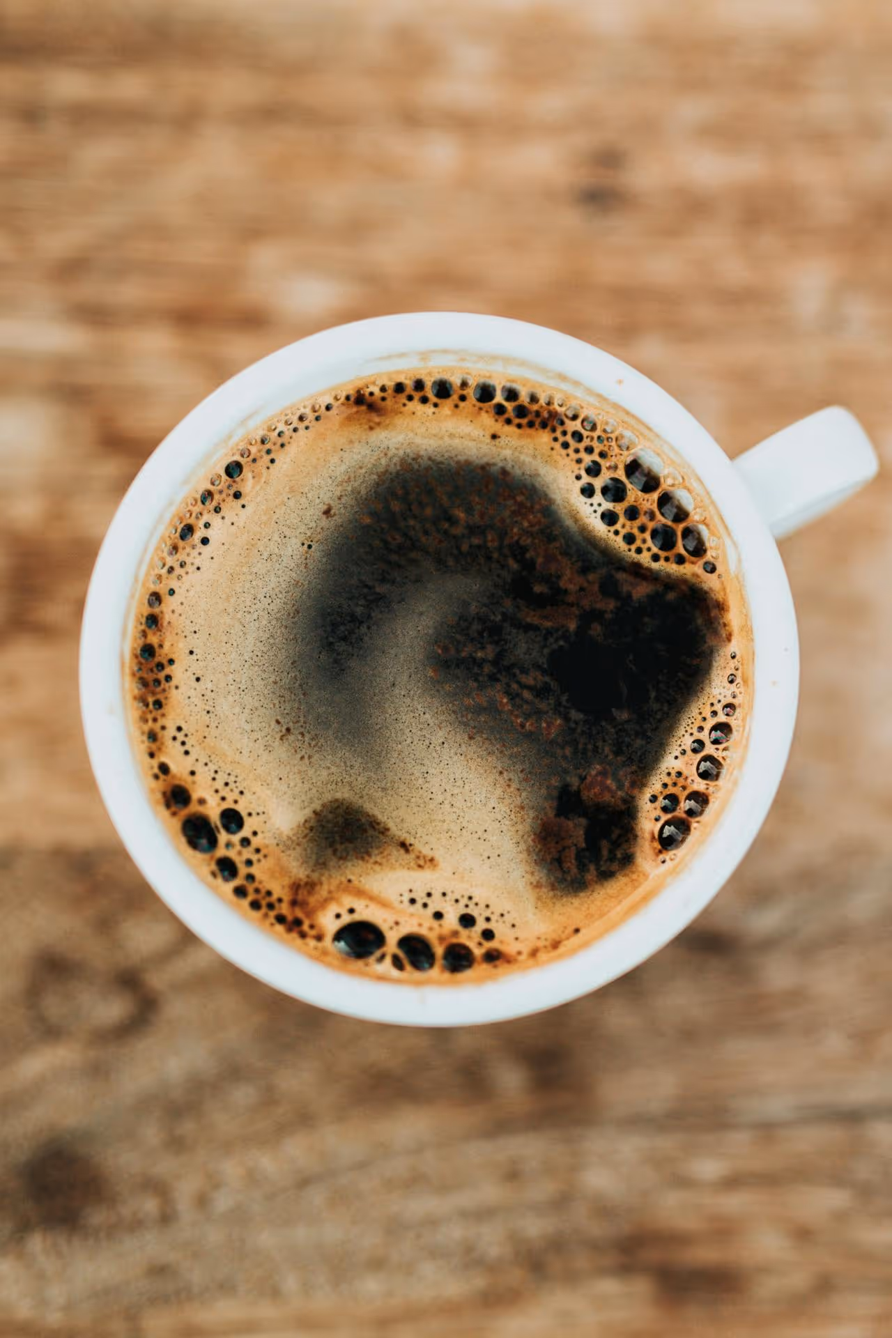 Close-up of a white cup filled with dark coffee, showcasing rich foam and bubbles against a rustic wooden background.