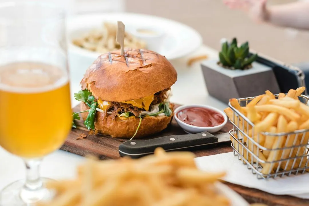 A juicy burger with melted cheese, cilantro, and sauce, served alongside crispy fries and a glass of golden beer on a wooden board.