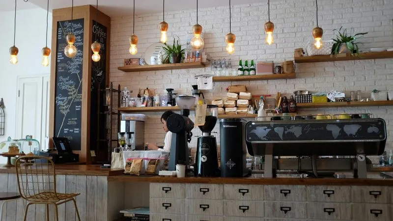 Cozy café interior with a rustic charm. Features hanging bulbs, chalkboard menu, coffee machines, plants on shelves, and wood counter with pastries. Relaxed ambiance.