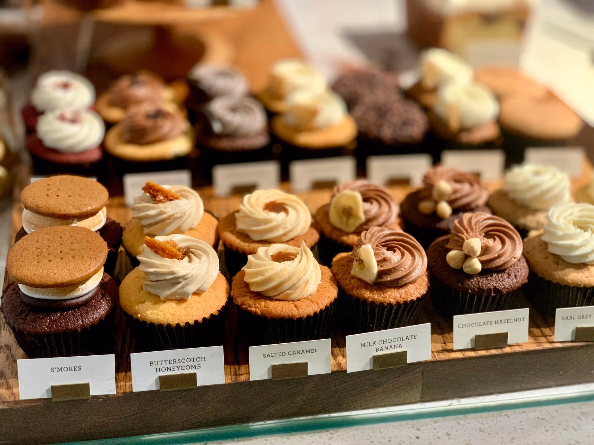 A variety of cupcakes displayed on a tray, each topped with frosting and decorations like cookies and nuts. Labels include flavors like S'mores and Salted Caramel.